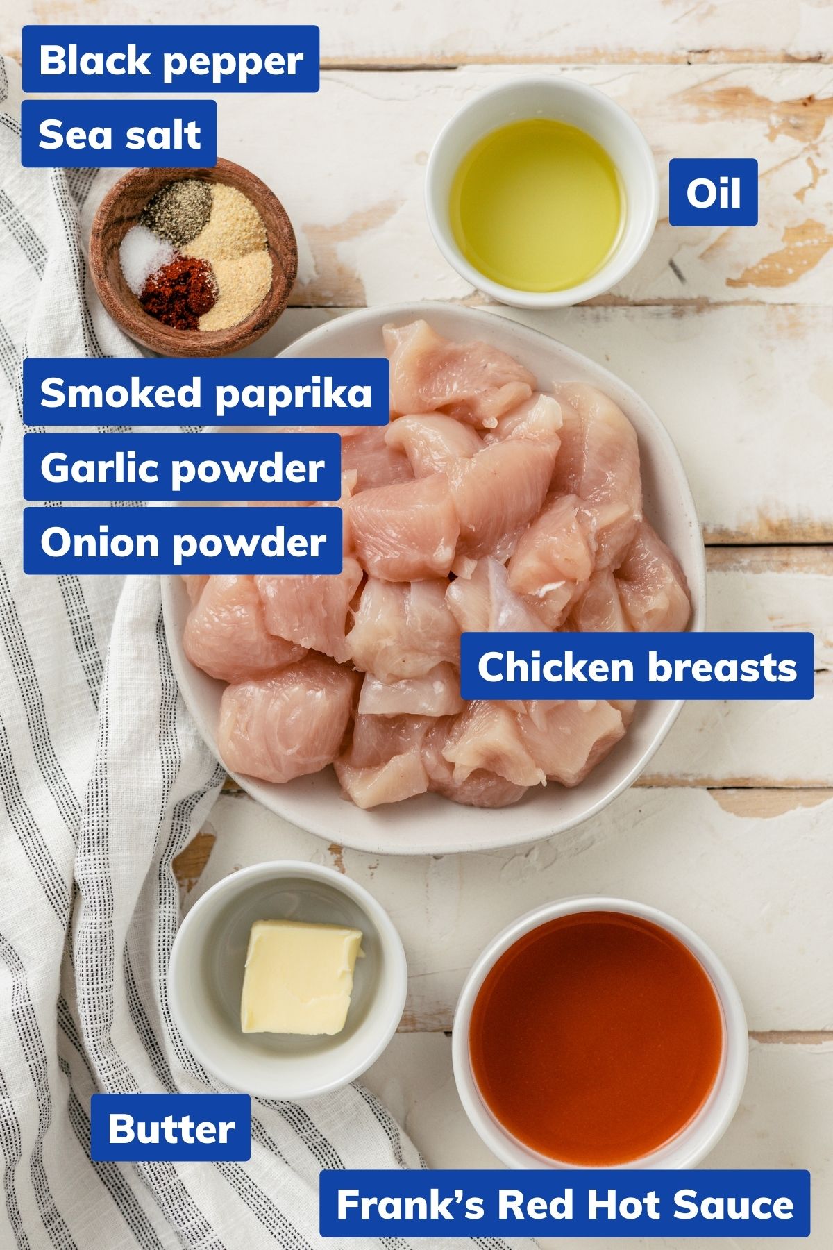 ingredients to make buffalo chicken bites (air fryer): Chicken breasts cut into bite-sized pieces, optional natural ancient sea salt, black pepper, smoked paprika, garlic powder, onion powder, a choice of avocado or olive oil, homemade buffalo sauce, Frank's Red Hot Sauce (or its buffalo wing variant), and unsalted butter are displayed in separate bowls.