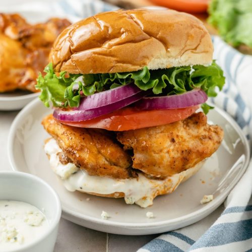 A tangy Buffalo Chicken Sandwich, with crispy chicken on a soft brioche bun, is garnished with butter lettuce, tomato, red onions, and drizzled with creamy blue cheese dressing on a plate.