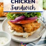 pinterest image of A tangy Buffalo Chicken Sandwich, with crispy chicken on a soft brioche bun, is garnished with butter lettuce, tomato, red onions, and drizzled with creamy blue cheese dressing on a plate.