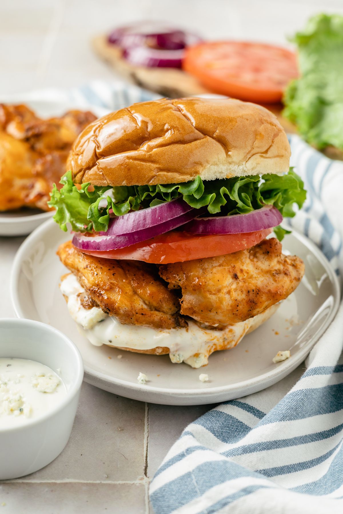 A tangy Buffalo Chicken Sandwich, with crispy chicken on a soft brioche bun, is garnished with butter lettuce, tomato, red onions, and drizzled with creamy blue cheese dressing on a plate.