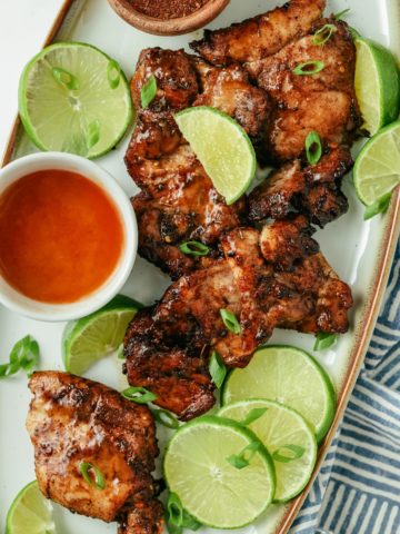 Juicy jerk chicken thighs, perfectly seasoned, surrounded by fresh lime slices, with a side of Sriracha and spicy jerk seasoning for added flavor