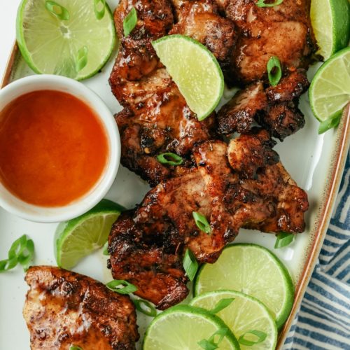 Juicy jerk chicken thighs, perfectly seasoned, surrounded by fresh lime slices, with a side of Sriracha and spicy jerk seasoning for added flavor