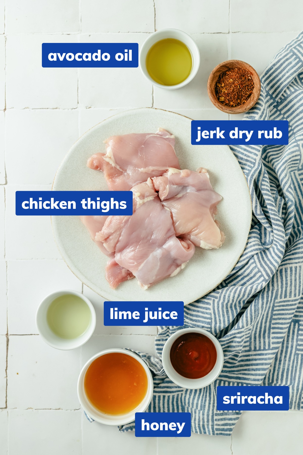 Ingredients for jerk chicken thighs, each in separate bowls: boneless, skinless chicken thighs, spicy jerk seasoning, and avocado oil, neatly arranged on a countertop. Also includes ingredients for a sauce in separate bowls: sriracha, honey, and lime juice